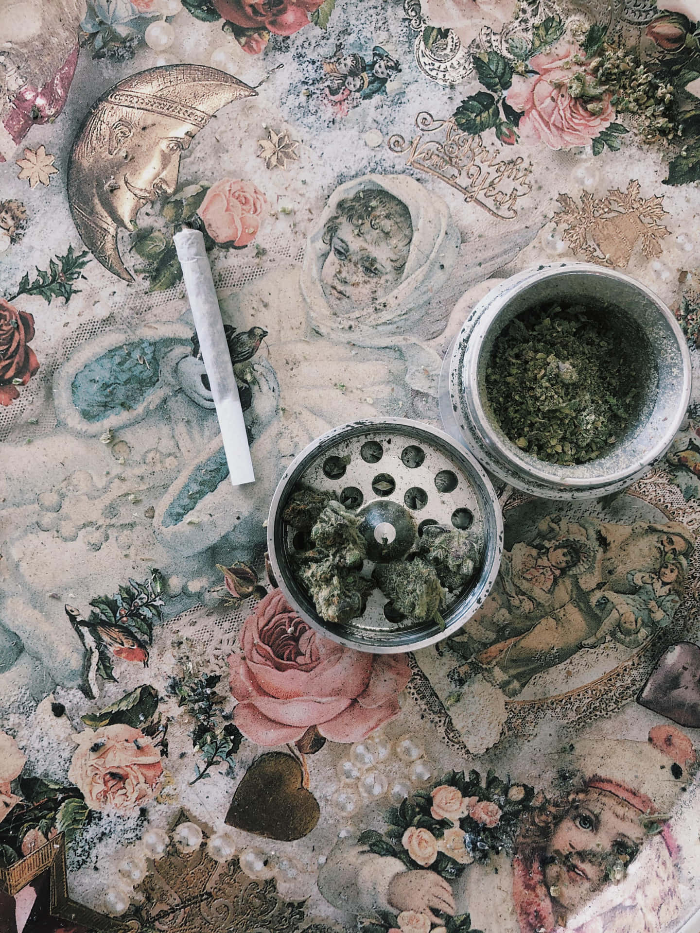 一支大麻煙和一個研磨器放在帶有小天使插圖的復古花卉桌布上。