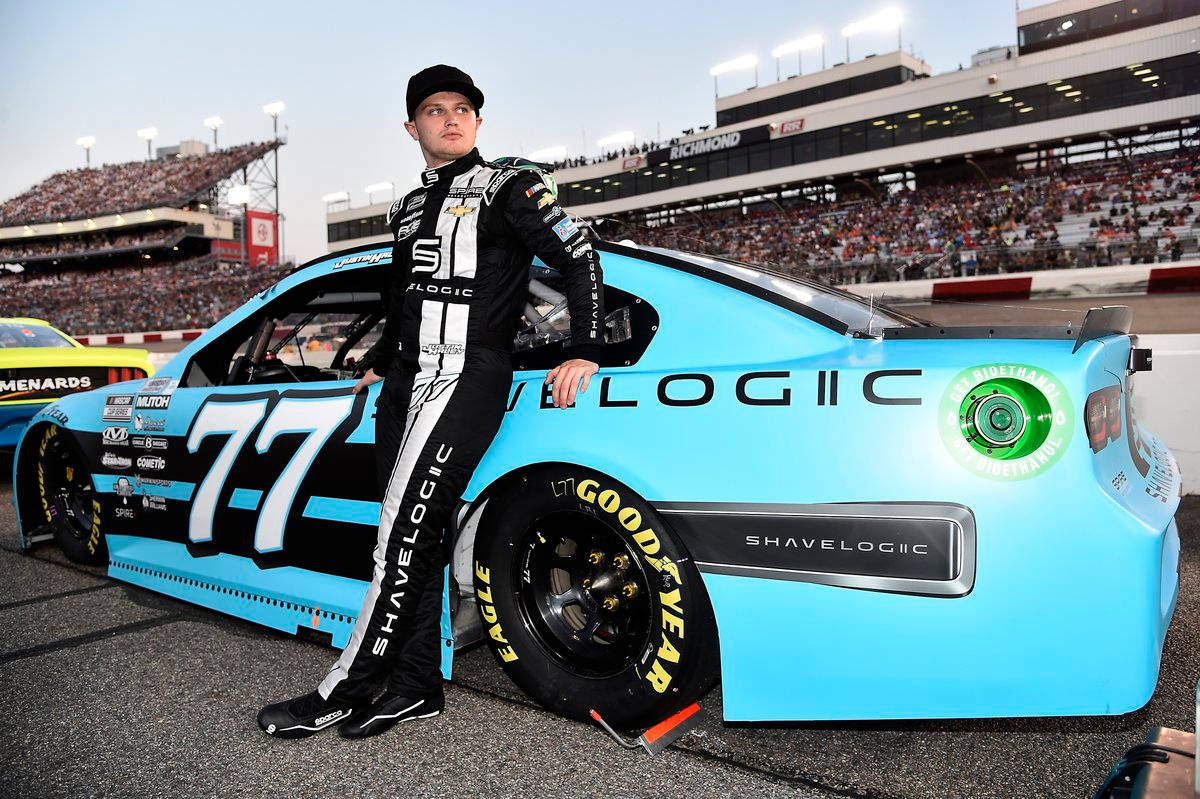 新星賈斯汀·黑利駕駛碧藍賽車 壁紙 — 一位身穿黑白裝備的NASCAR車手在里士滿賽車場靠著他的淺藍色#77 ShaveLogic賽車，身後是擠滿觀眾的看台。