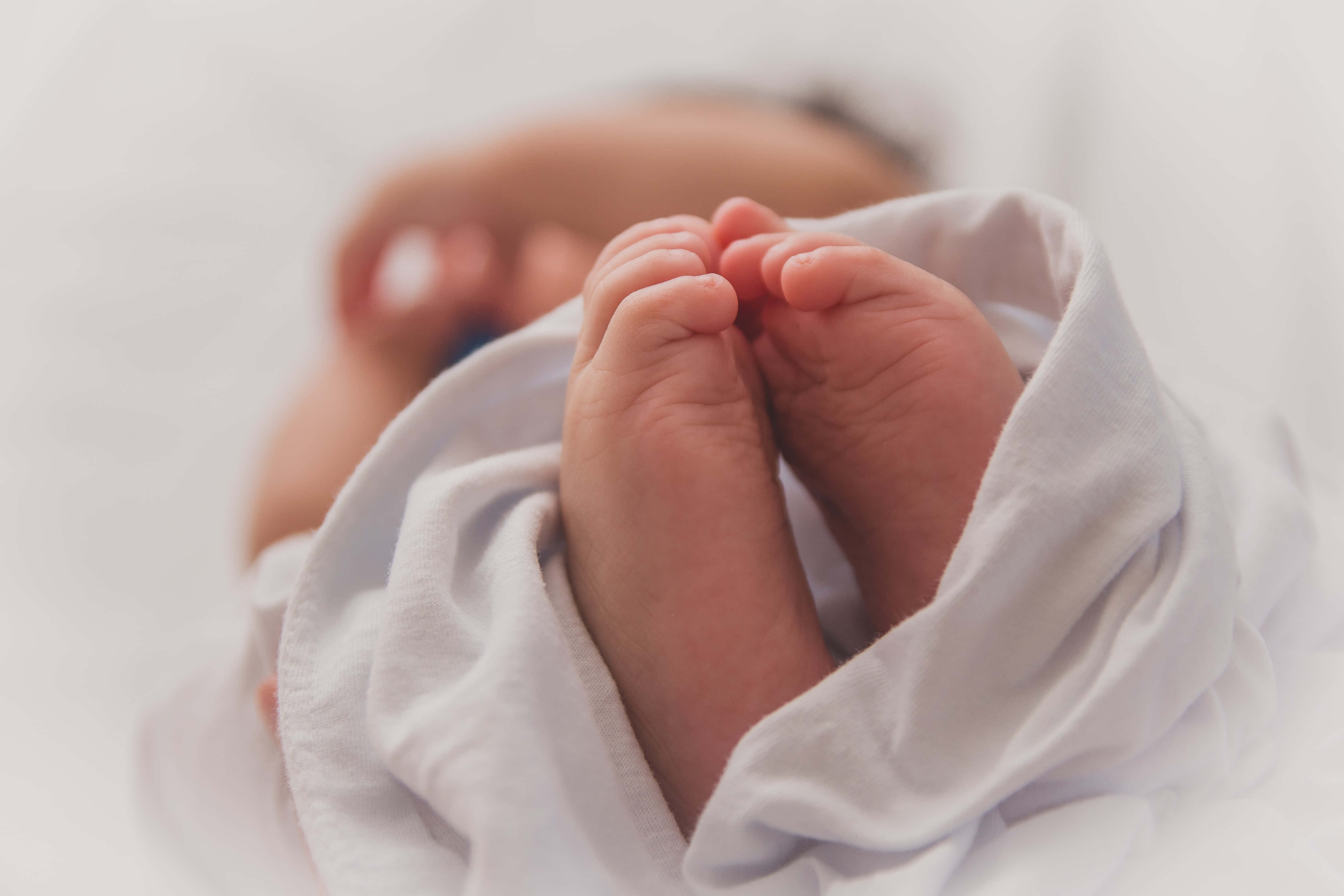 Baby Boy 桌布 壁紙 — 新生兒小腳的特寫，包裹在柔軟的白色布料中，背景有模糊的臉部。