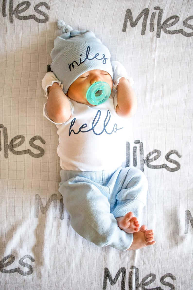 新生男嬰文字服裝照片 壁紙 — 一個名叫Miles的熟睡新生嬰兒，戴著印有他名字和「你好」字樣的配套帽子和襯衫，躺在圖案毯子上休息。