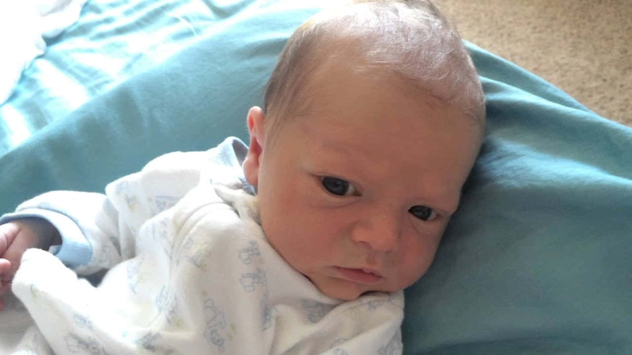 新生男嬰的幸福時光 壁紙 — 一個深色眼睛的新生嬰兒，包裹在白色圖案連體衣中，安詳地躺在青綠色毯子上。