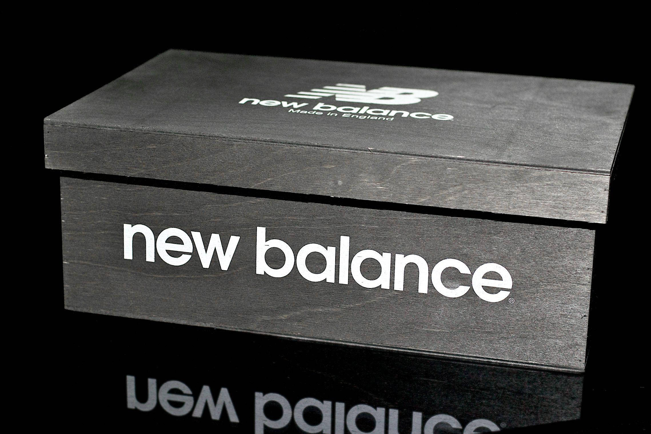 新百倫鞋盒 壁紙 — 一個時尚的黑色New Balance鞋盒，帶有白色品牌標誌，放置在深色背景的反射表面上。