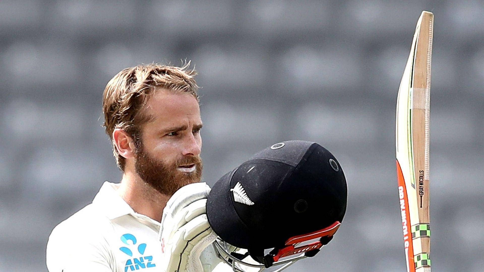 新西蘭板球 Kane Williamson 壁紙 — 一位專注的板球運動員身穿白色制服，在模糊的體育場座位前手持頭盔和球棒。