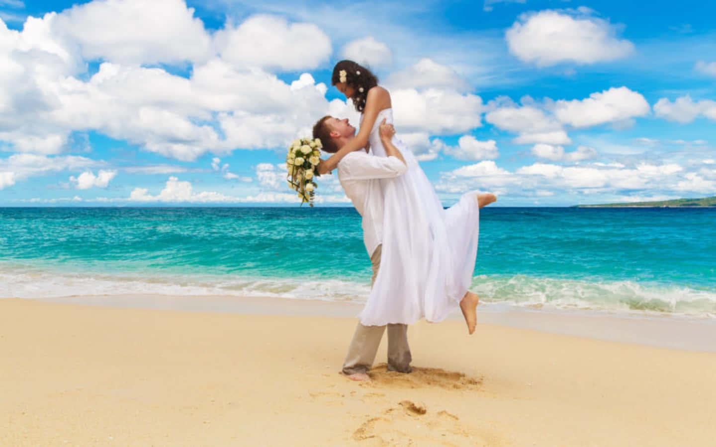 Wedding Couple 桌布 壁紙 — 新郎愉快地抱起穿著白色婚紗、手持花束的新娘，背景是陽光明媚的海灘、碧綠的海洋和蓬鬆的雲朵。