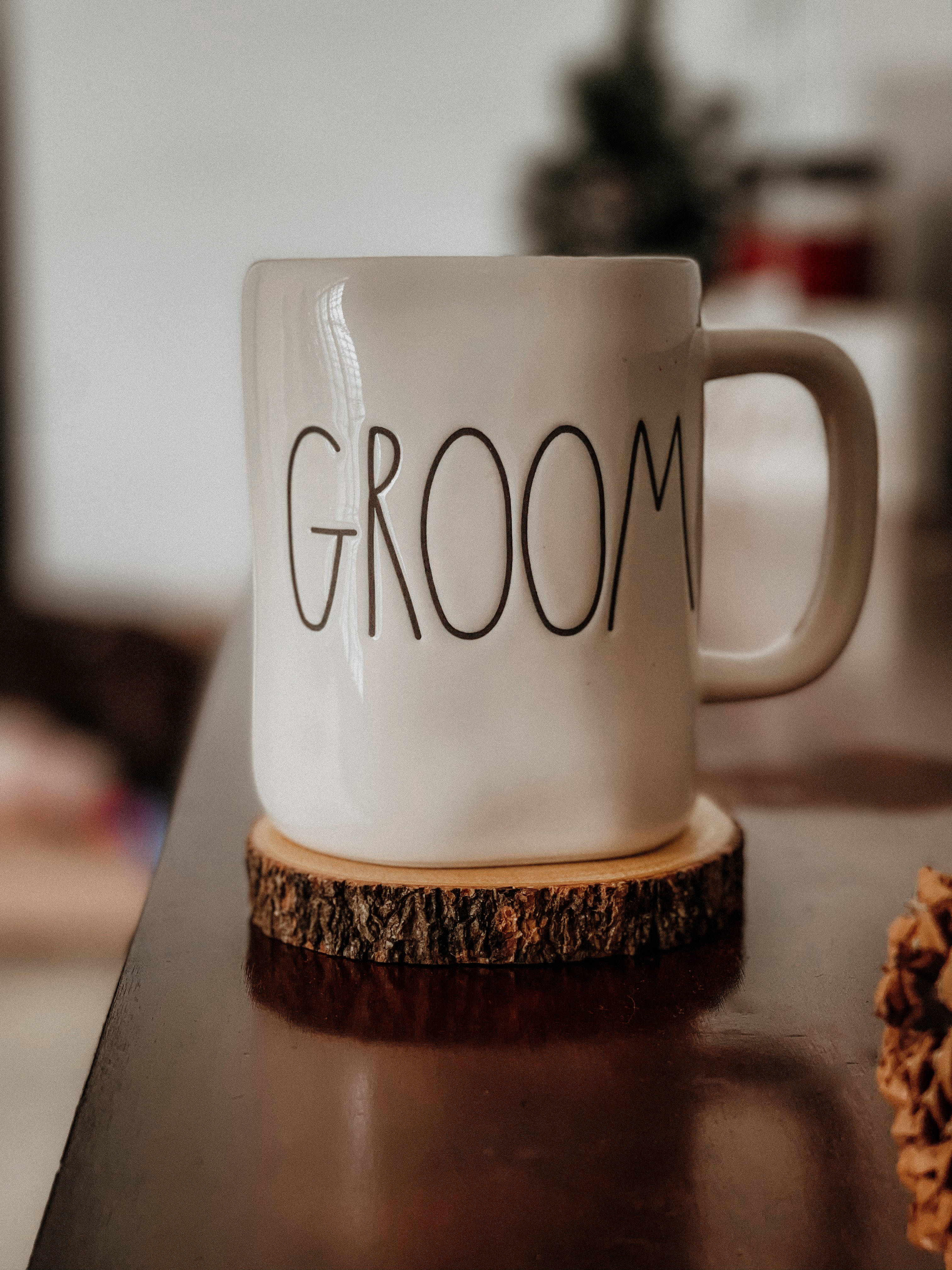 新郎的白瓷杯 壁紙 — 一個白色陶瓷杯，上面標有「GROOM」，放在深色桌子上的質樸木杯墊上，背景柔和模糊。