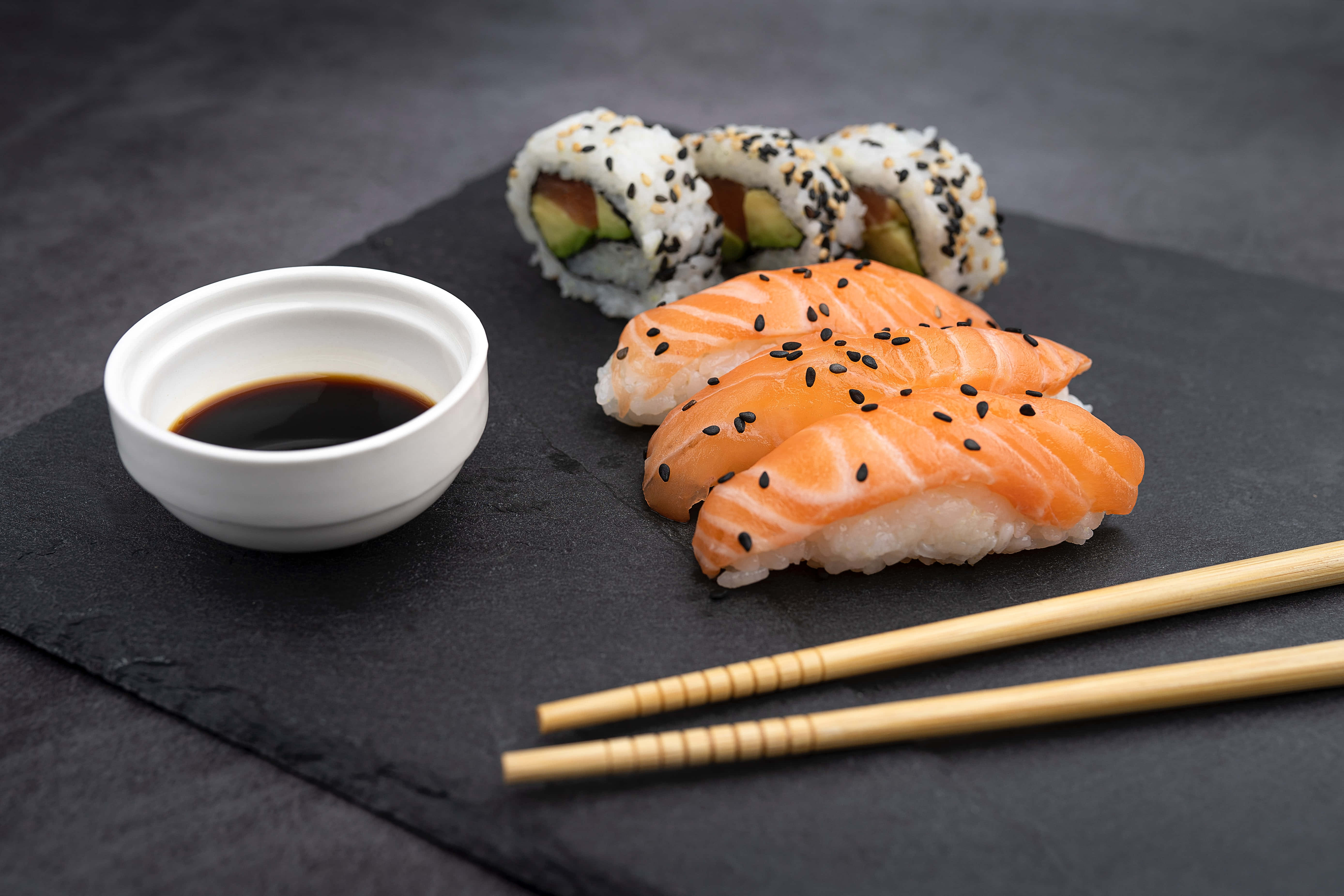 Sushi 桌布 壁紙 — 新鮮三文魚握壽司和牛油果卷，飾以黑芝麻，旁邊有醬油和筷子，放在深色石板盤上。