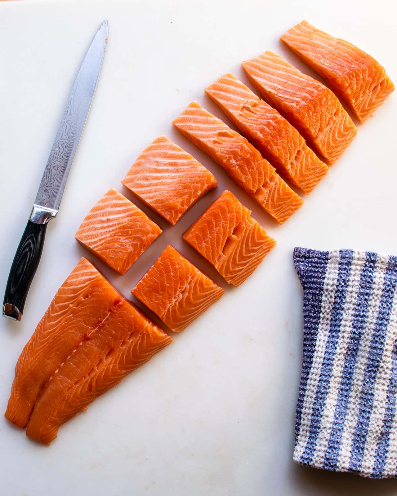 Salmon 桌布 壁紙 — 新鮮三文魚柳切成塊，放在白色砧板上，旁邊有刀和條紋布。