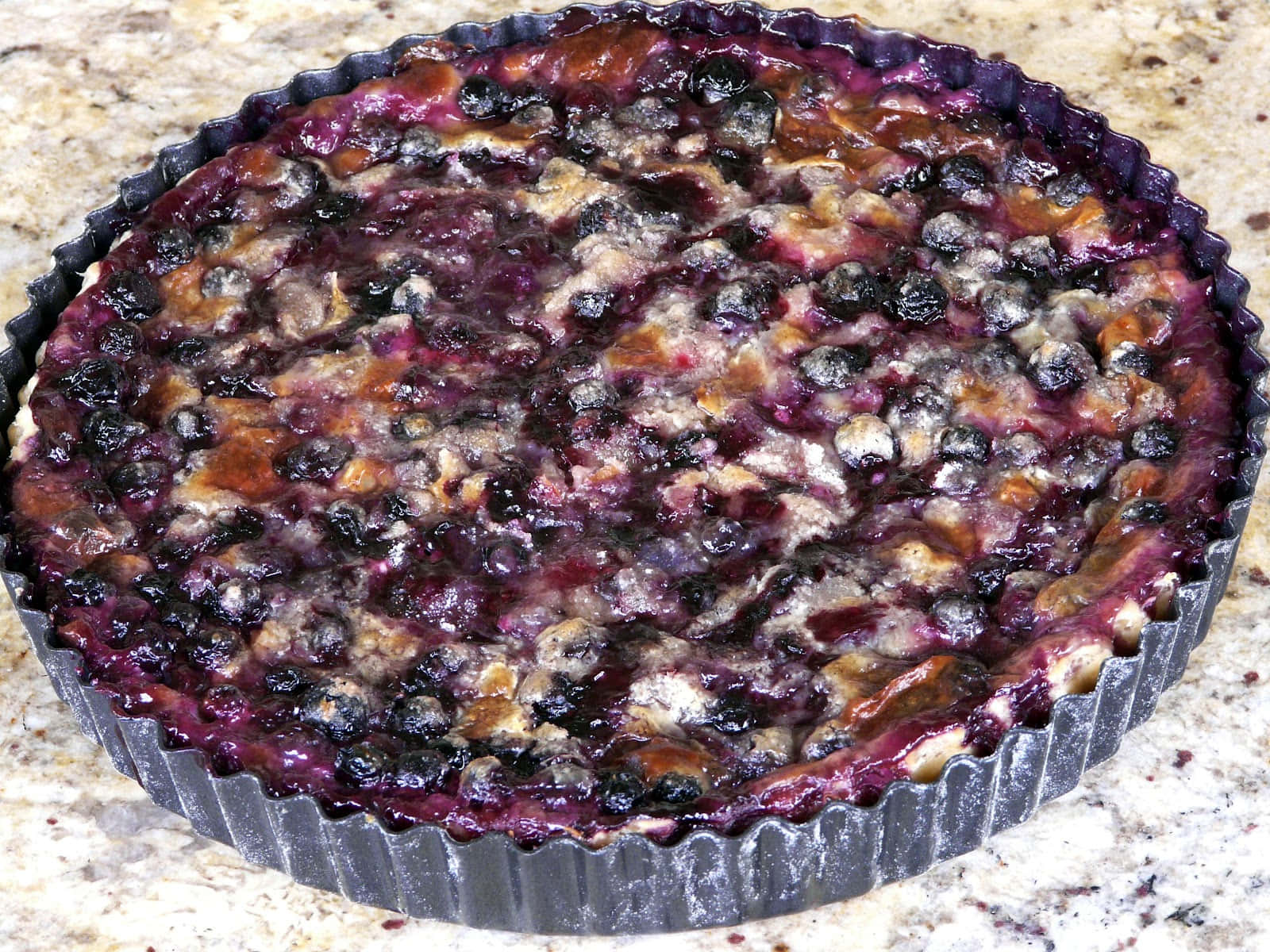 Blueberries Tart 桌布 壁紙 — 新鮮出爐的藍莓撻，多汁的莓果和金黃色的外皮，放在一個有凹槽的金屬烤盤中，置於斑點點的工作檯面上。