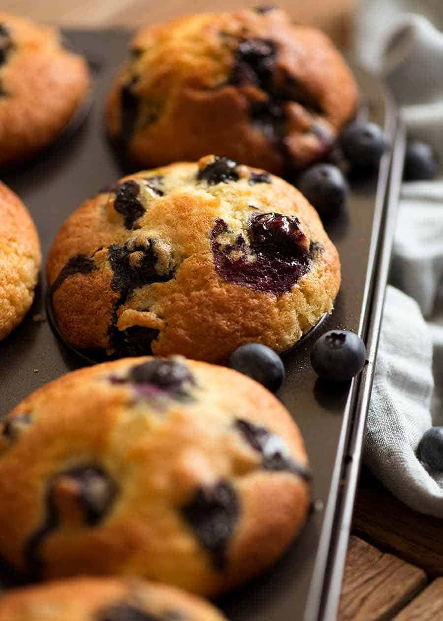 Blueberry Muffins 桌布 壁紙 — 新鮮出爐的藍莓鬆餅，金黃外皮，多汁莓果，放在深色托盤上，旁邊有一塊亞麻布。