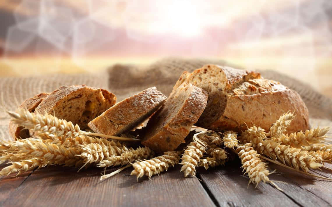 Bakery 桌布 壁紙 — 新鮮出爐的鄉村麵包片和金黃色麥穗放在一張飽經風霜的木桌上，沐浴在溫暖的陽光中。