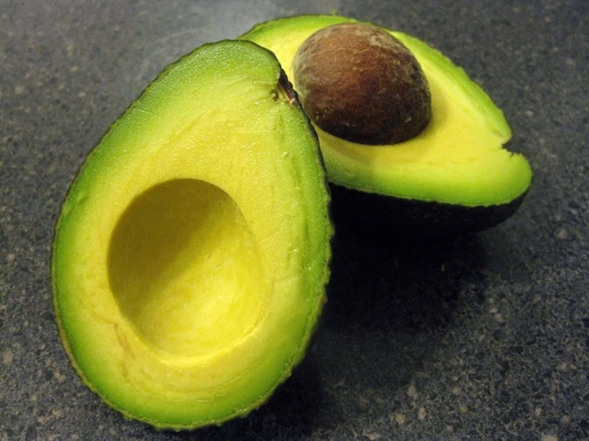 Avocado 桌布 壁紙 — 新鮮切半的牛油果露出其奶油黃色的果肉和深色的果核，放在有斑點的枱面上。