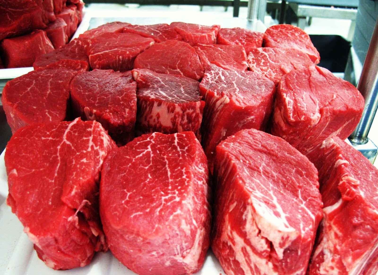 meat 桌布 壁紙 — 新鮮切好的牛肉扒，呈現豐富的紅色和白色大理石花紋，放在白色托盤上，在市場環境中展示。