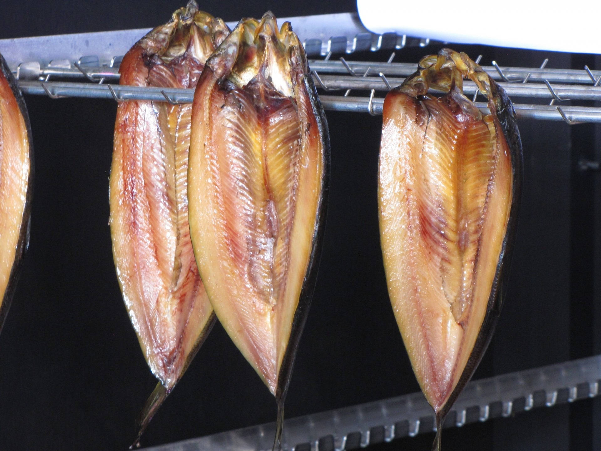 新鮮去骨鯡魚 壁紙 — 切片魚掛在金屬架上晾乾，背景黑暗，展現細膩質感和豐富琥珀色調。