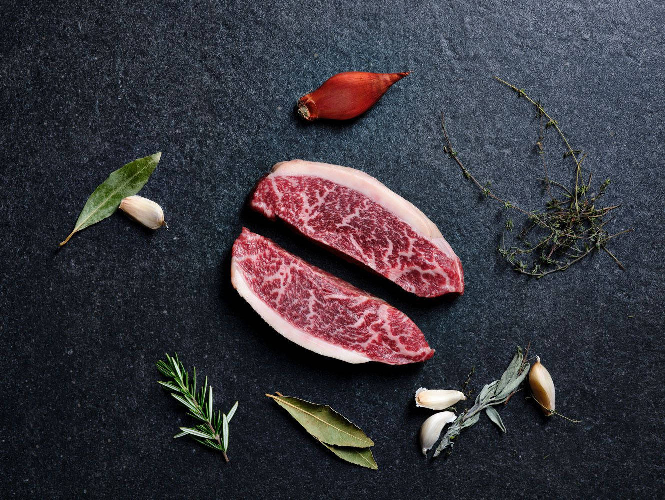 兩塊生牛肉扒配香草和大蒜，放在深色石板背景上。