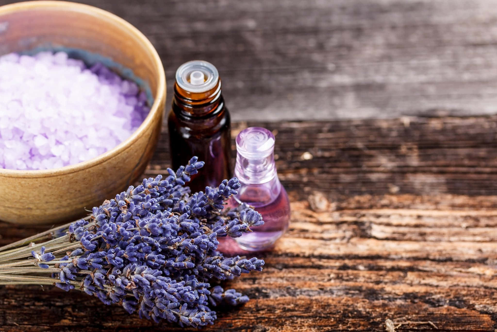Essential Oil 桌布 壁紙 — 新鮮嘅薰衣草枝條放喺一個裝滿紫色浴鹽嘅木碗同玻璃樽精油旁邊，喺一塊樸實風化嘅木表面上。