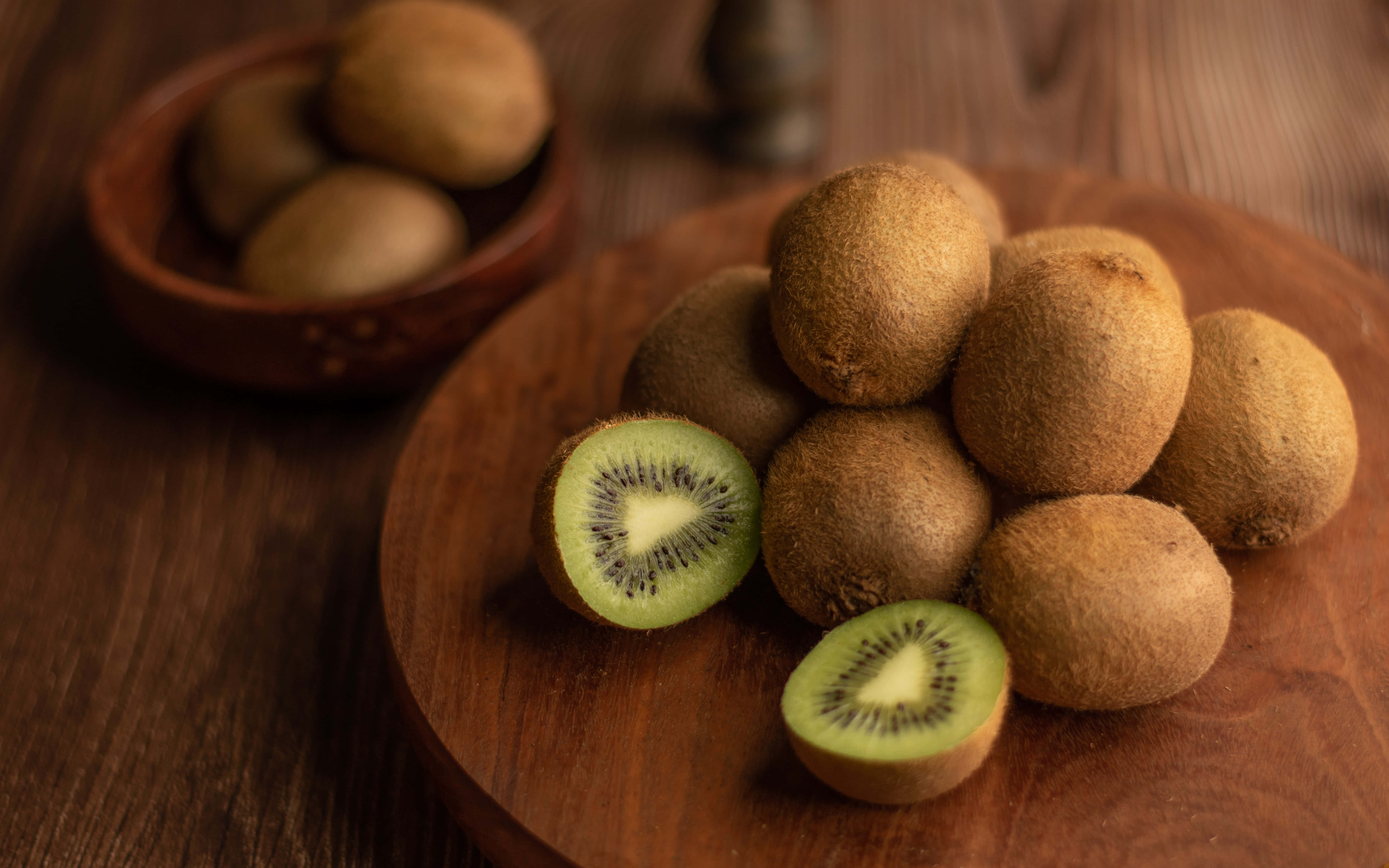 Kiwi 桌布 壁紙 — 新鮮奇異果，帶有絨毛棕色外皮和鮮綠色果肉，放在溫暖的木板上，背景有一個小碗。