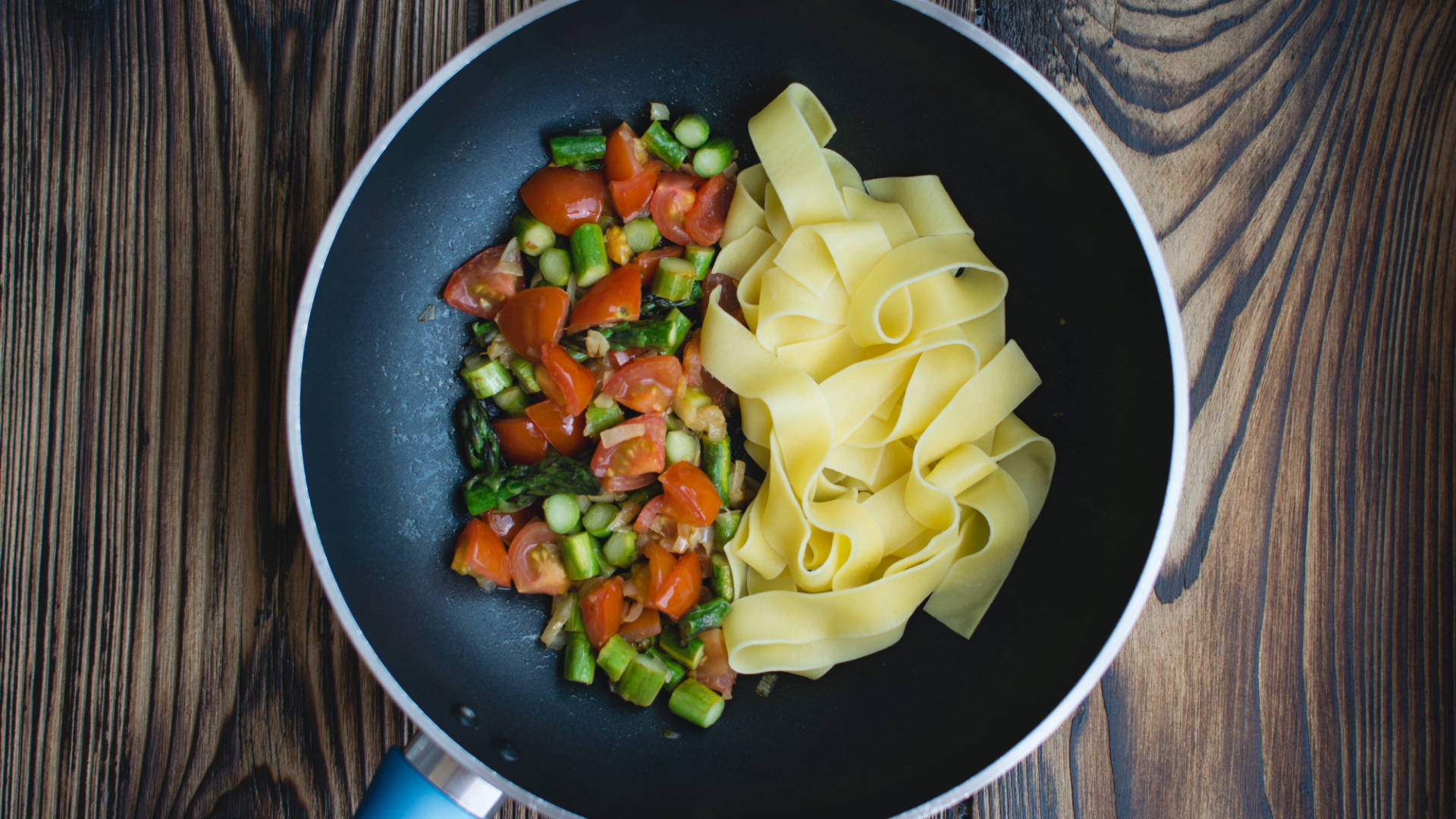 Pasta 桌布 壁紙 — 新鮮意粉和色彩繽紛的炒蔬菜並排放在黑色煎鍋中，置於鄉村風格的木質表面上。