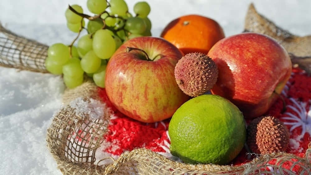 Winter Fruits 桌布 壁紙 — 新鮮水果包括蘋果、葡萄、荔枝和青檸，放在雪旁的一塊紅色圖案布上。