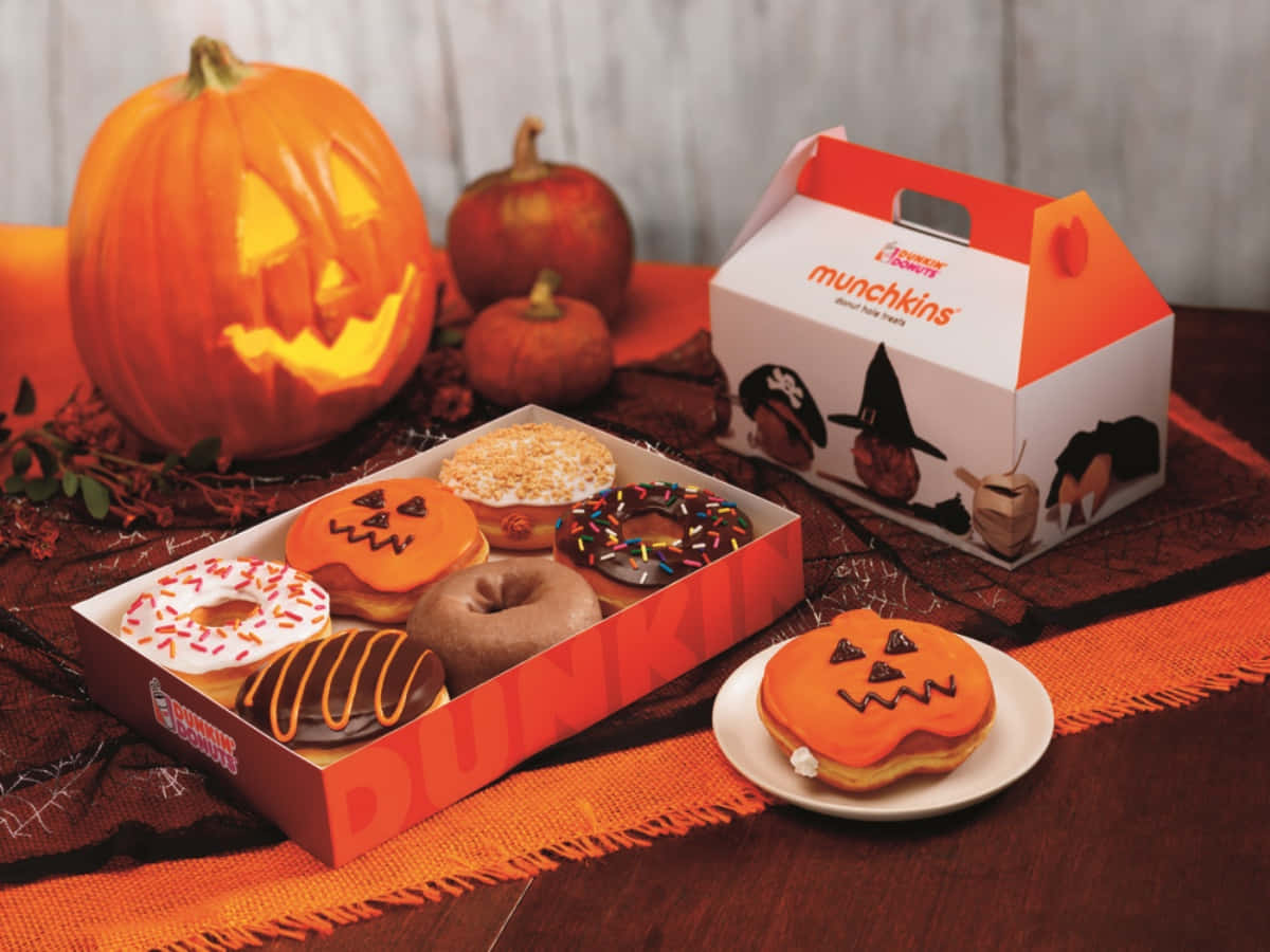 新鮮沖煮的咖啡與甜甜圈，來自Dunkin' Donuts！ 壁紙 — 萬聖節主題冬甩，帶有傑克南瓜燈面孔，旁邊放著發光雕刻南瓜和節日包裝，置於橙色布上。