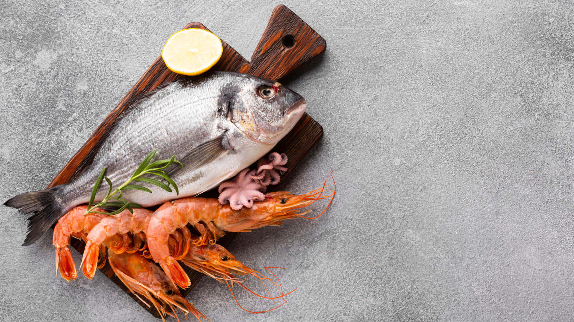 Seafood 桌布 壁紙 — 新鮮海鱸魚、蝦和八爪魚擺放在木板上，配以檸檬和迷迭香，置於灰色石面上。