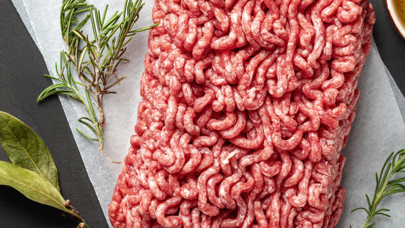 Red Meat 桌布 壁紙 — 新鮮生免治牛肉放在牛油紙上，旁邊有迷迭香枝和月桂葉，置於深色石板表面上。