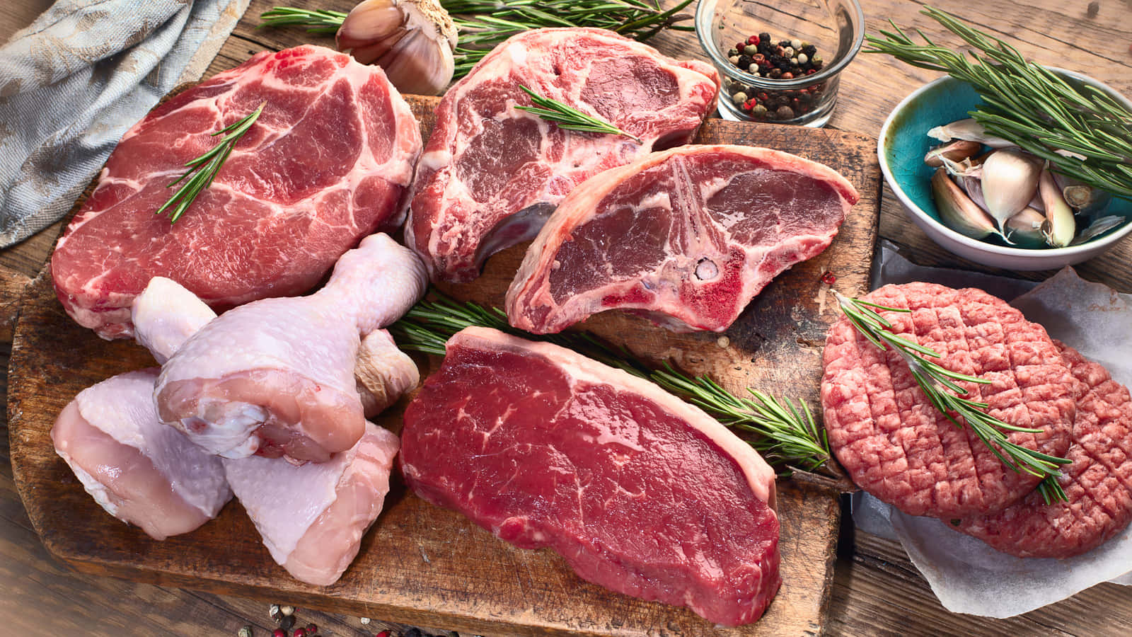 Red Meat 桌布 壁紙 — 新鮮生肉包括牛扒、雞大腿和漢堡扒，連同香草和大蒜擺放在質樸的木砧板上。