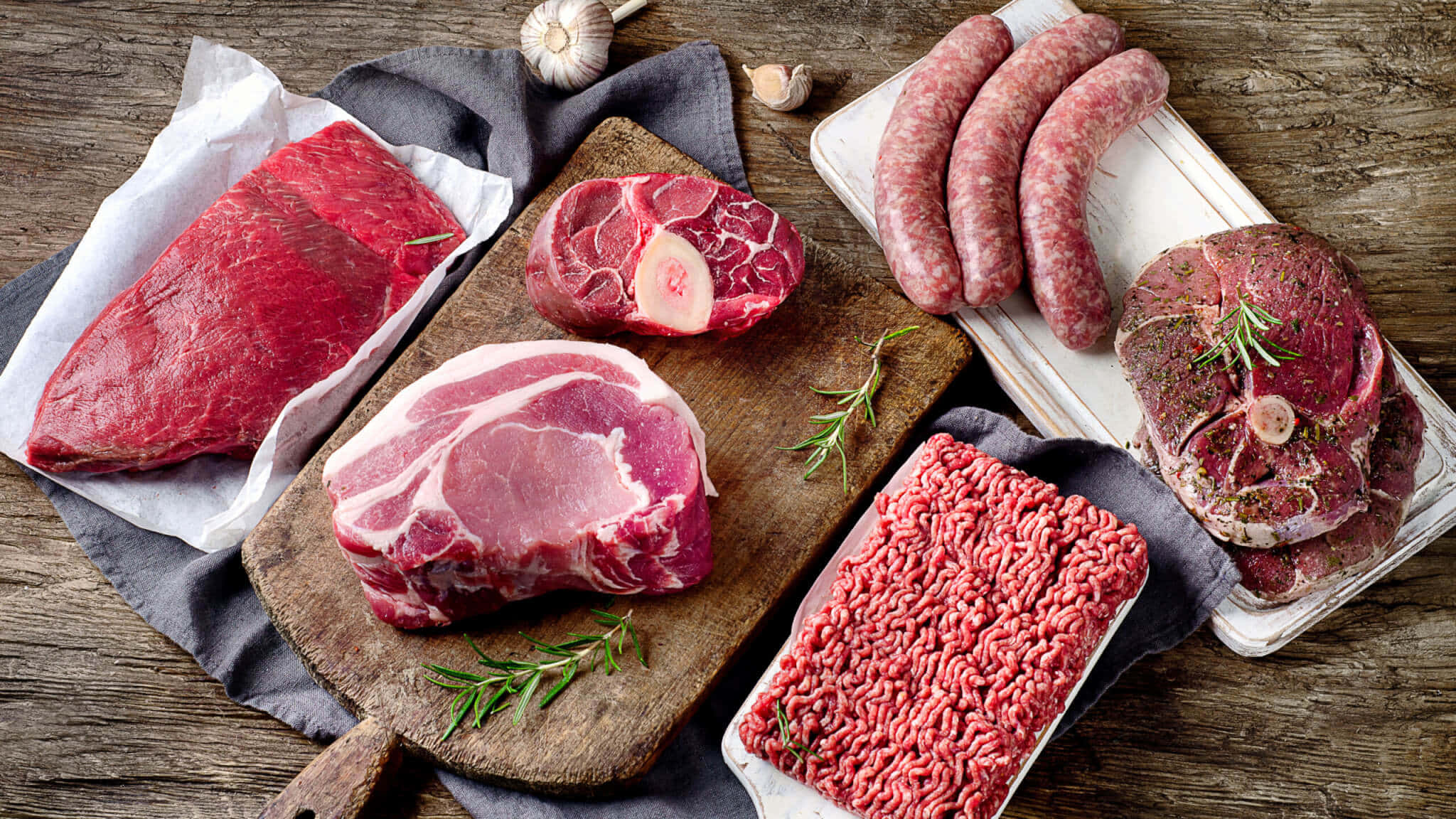 Red Meat 桌布 壁紙 — 新鮮生肉包括牛扒、香腸和免治牛肉，放在木板上，配以迷迭香和大蒜，置於鄉村風木桌上。
