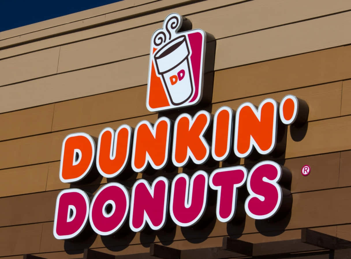 新鮮的Dunkin' Donuts，您最愛的開晨方式 壁紙 — 一個鮮橙色和粉紅色的Dunkin' Donuts標誌，帶有咖啡杯標誌，安裝在米色建築物外牆上，背景是晴朗的藍天。