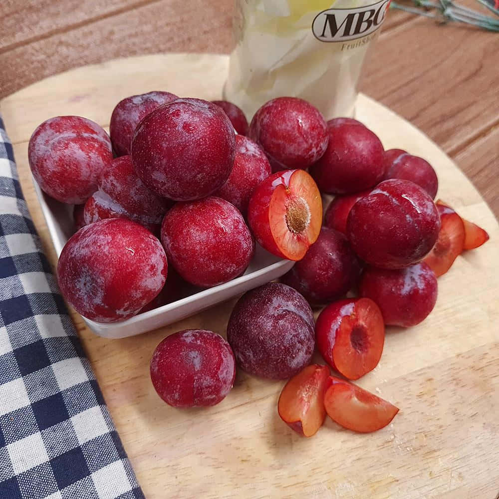 Red Plum 桌布 壁紙 — 新鮮的紅李子帶有粉狀果粉，放在白色碟子中，置於木板上，旁邊有格仔布和一杯飲品。