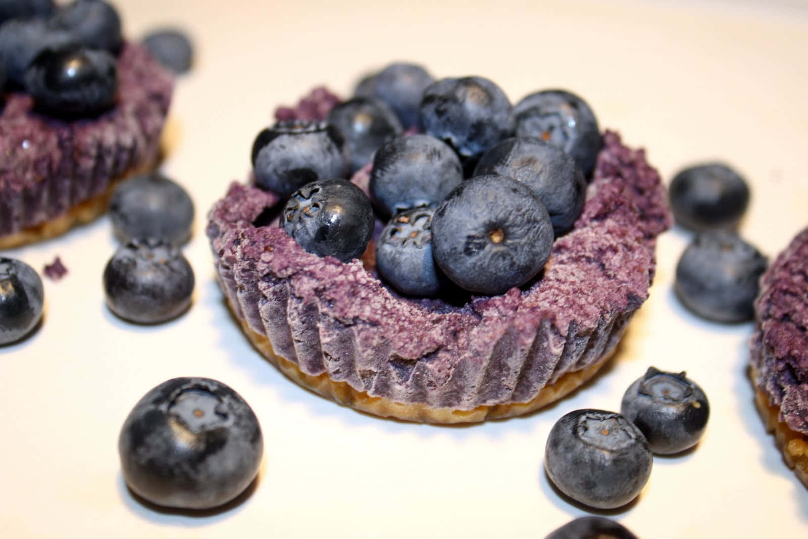 Blueberries Tart 桌布 壁紙 — 新鮮藍莓撻的微距特寫，帶有紫色餡料和散落的莓果，置於白色表面上。