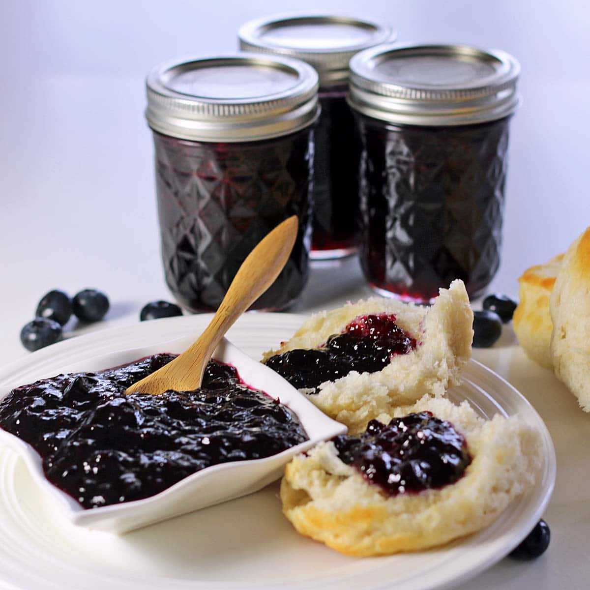 Blueberry Jam 桌布 壁紙 — 新鮮藍莓醬裝在玻璃罐中，配上金黃色餅乾，上面塗有果醬，放在白色盤子上。