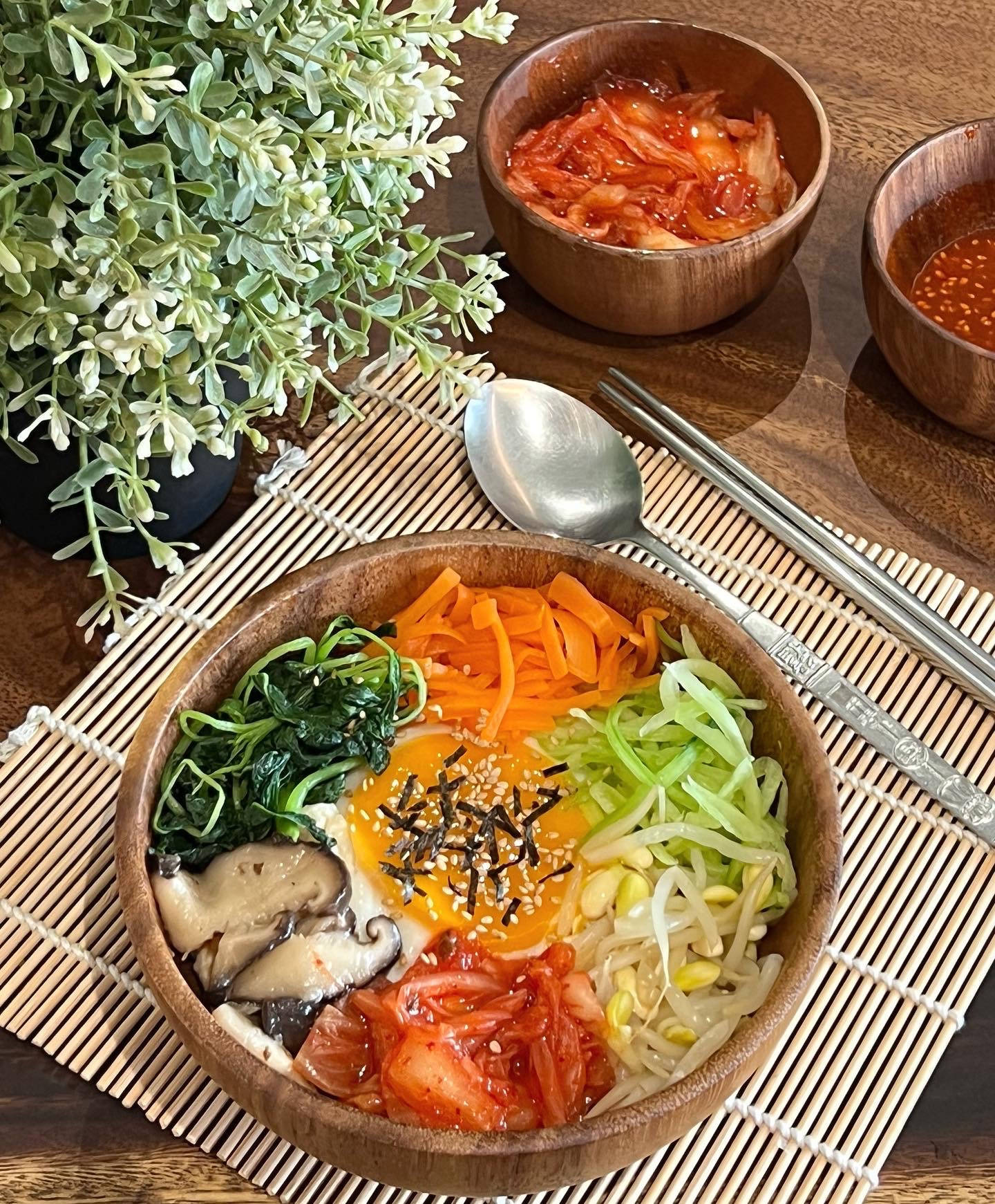 新鮮食材製作的韓式拌飯 壁紙 — 一碗色彩繽紛的韓國石鍋拌飯，配上各色蔬菜、芝麻和太陽蛋，放在竹蓆上，旁邊有泡菜和筷子。