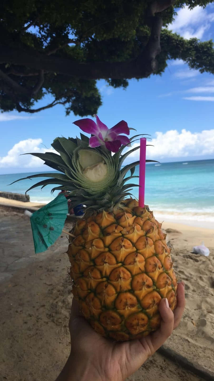 手在沙灘上拿著菠蘿飲品、蘭花和吸管。