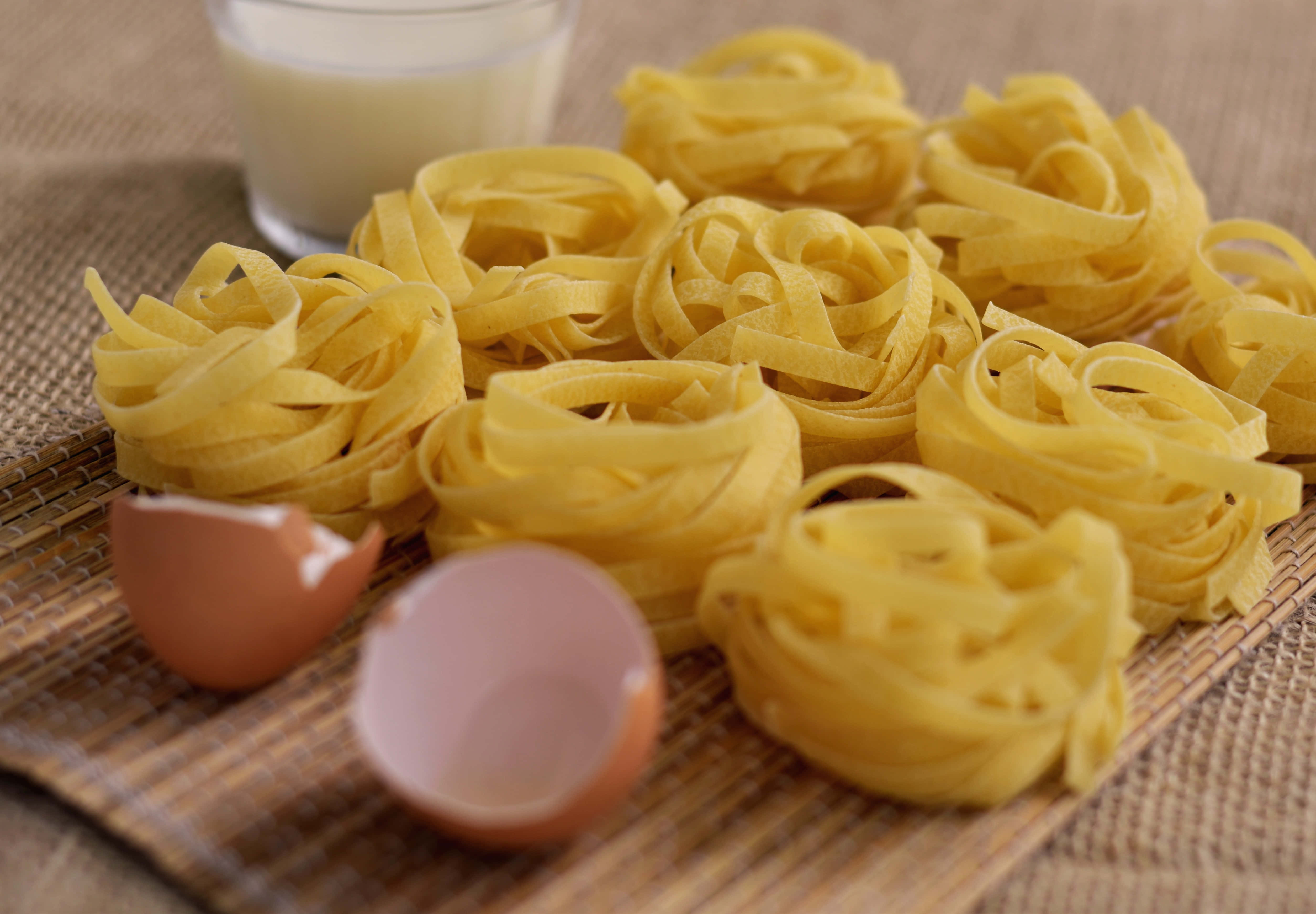 Brown Eggs 桌布 壁紙 — 新鮮黃色意粉巢排列在編織墊上，旁邊有破碎的蛋殼和一杯牛奶。