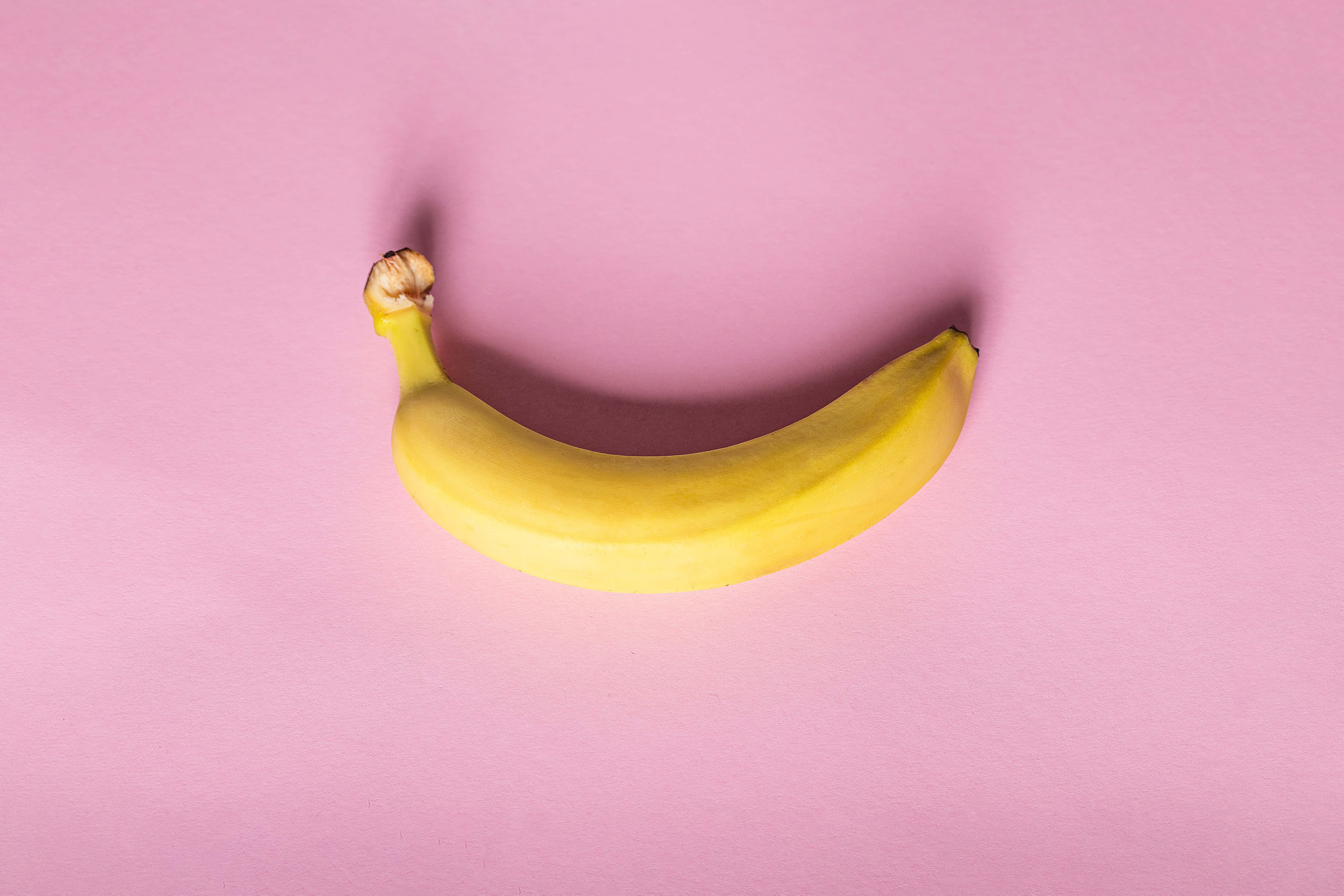 新鮮黃色香蕉，即刻享用 壁紙 — 一根成熟的黃色香蕉在柔和的粉紅色背景上優雅彎曲，帶有微妙的陰影。