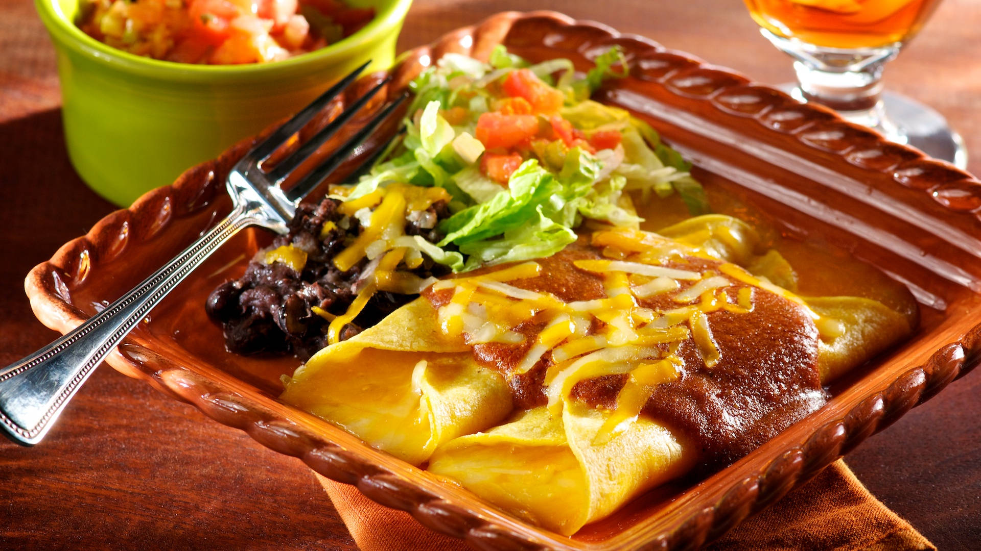 方木盤裝 enchiladas 壁紙 — 一盤冒著熱氣的玉米捲餅，淋上紅醬和融化的芝士，旁邊放著黑豆和新鮮的墨西哥辣醬，擺在質樸的木桌上。