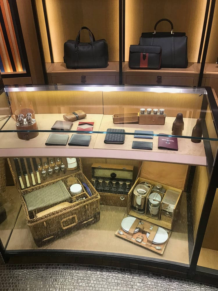 於 Moynat London 店內盡情購物 壁紙 — 豪華皮革製品和復古野餐套裝陳列在溫暖照明的玻璃展示櫃中，配有木質層架。