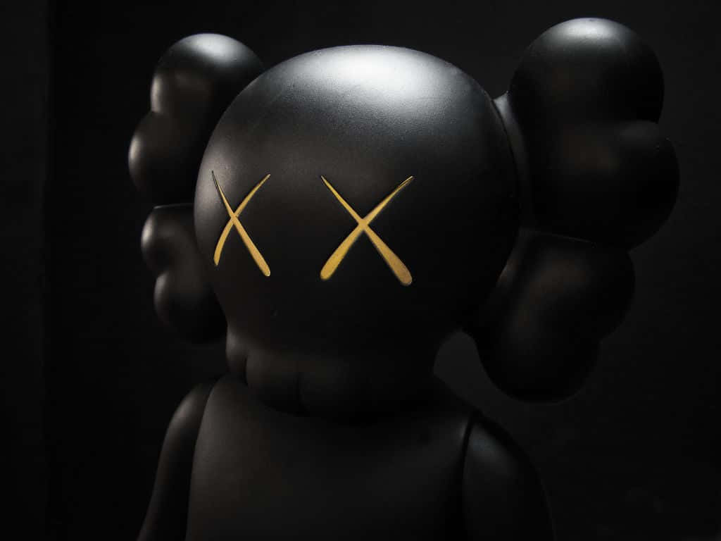 於 Wallpapers.com 購買原創 KAWS 藝術品 壁紙 — 一個光澤的黑色KAWS雕塑，帶有金色X眼睛，站在深色背景前，其標誌性的氣球狀耳朵在聚焦燈光下微微發光。