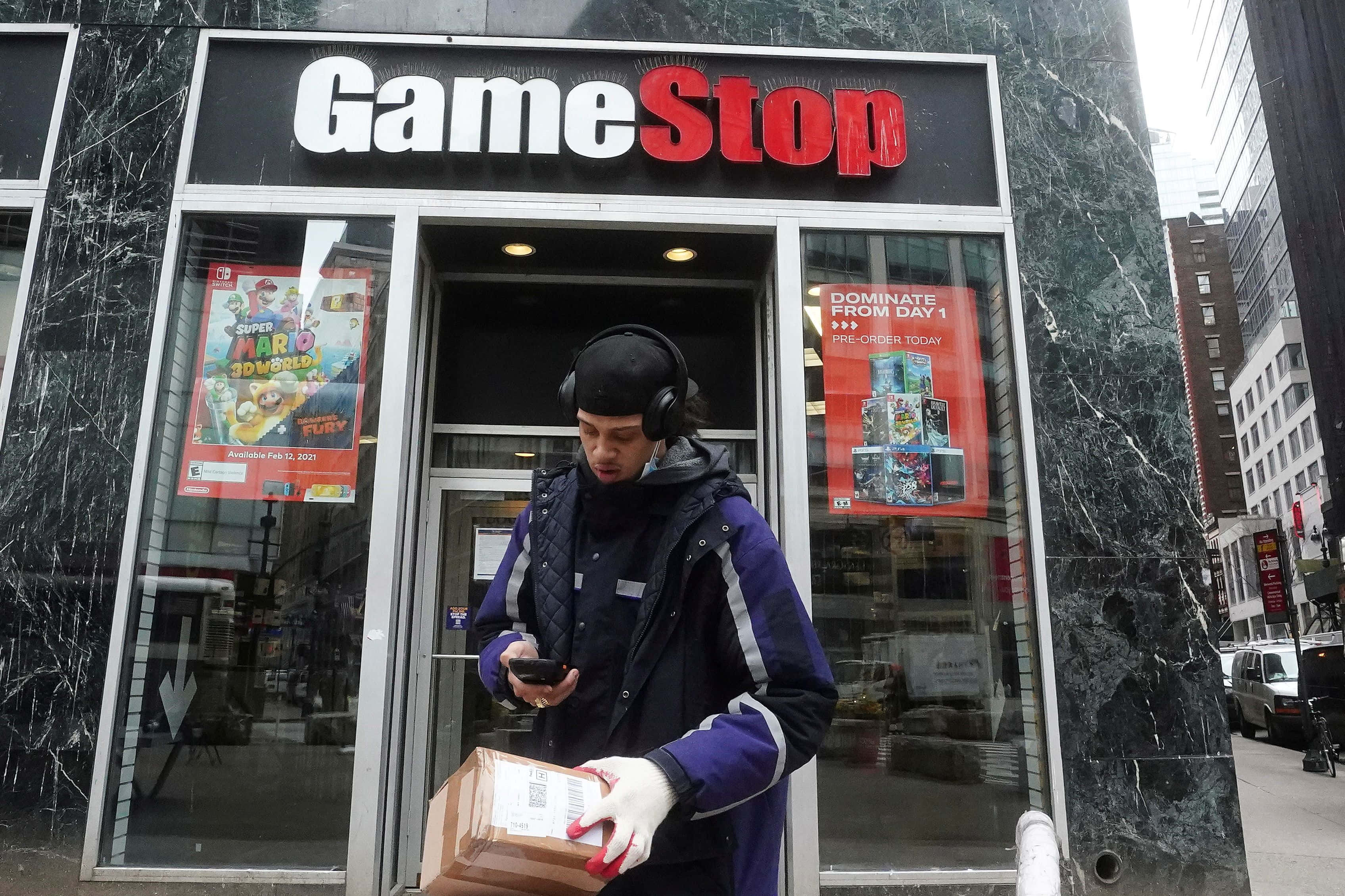 於Gamestop尋找你的終極遊戲體驗 壁紙 — 一名身穿紫色外套、戴著耳機的送貨員站在GameStop店外，手持包裹並查看手機。