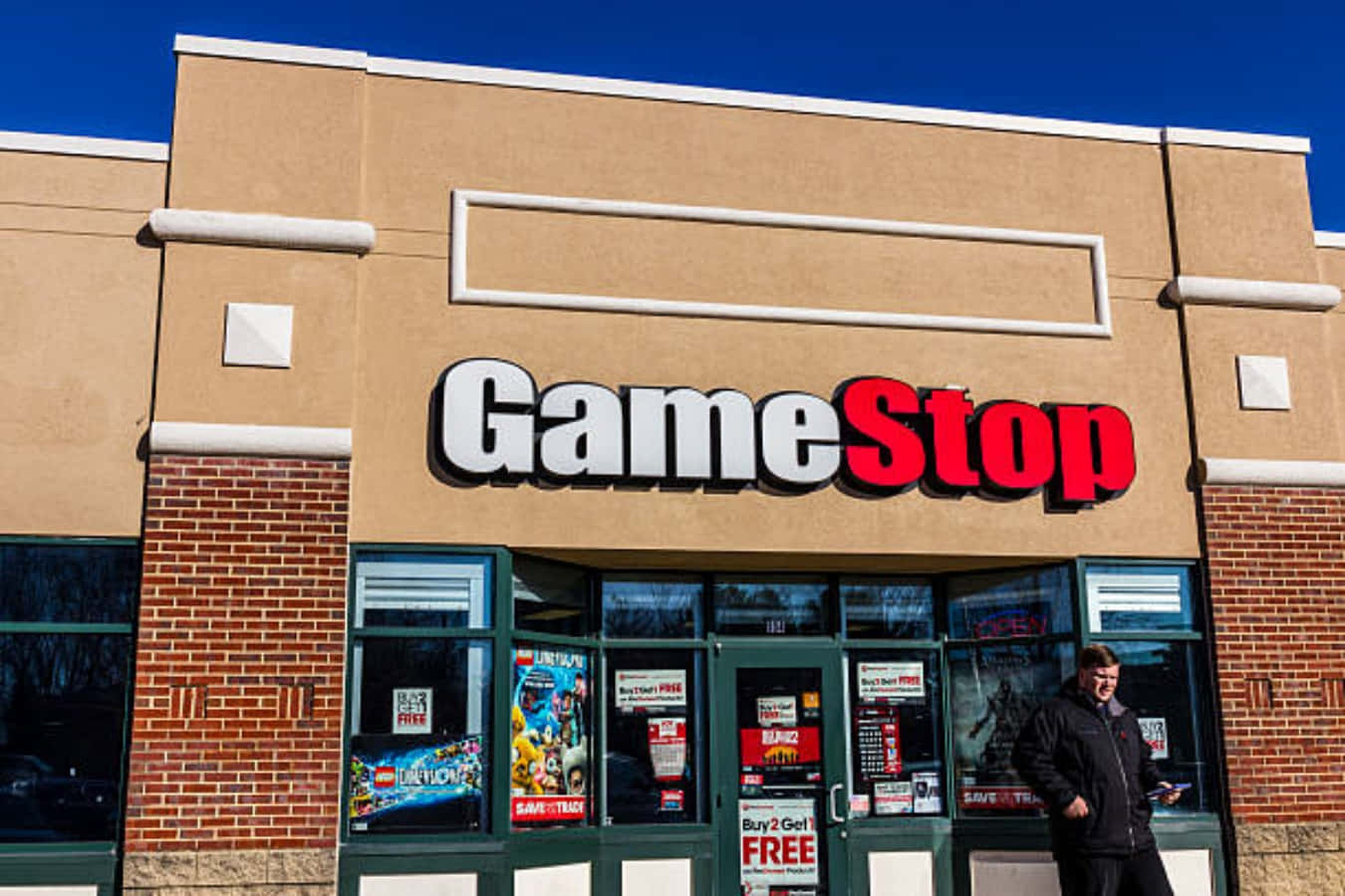 於Gamestop購買遊戲裝備！ 壁紙 — 晴朗陽光普照的日子，在GameStop商店外，一名男子站在清澈的藍天下。