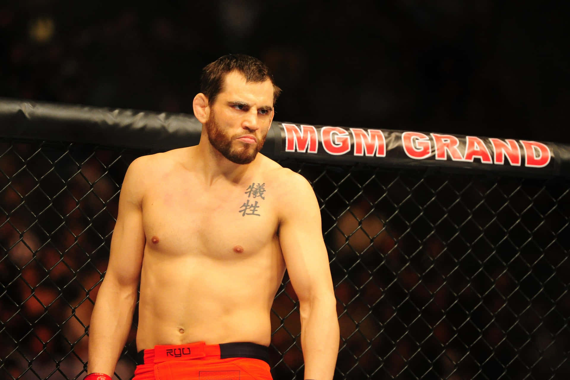 於MGM Grand Garden Arena的強悍UFC選手約恩·菲奇 壁紙 — 一位留鬍鬚、胸前有紋身的肌肉戰士，在MGM Grand的場館燈光下站在MMA籠內。