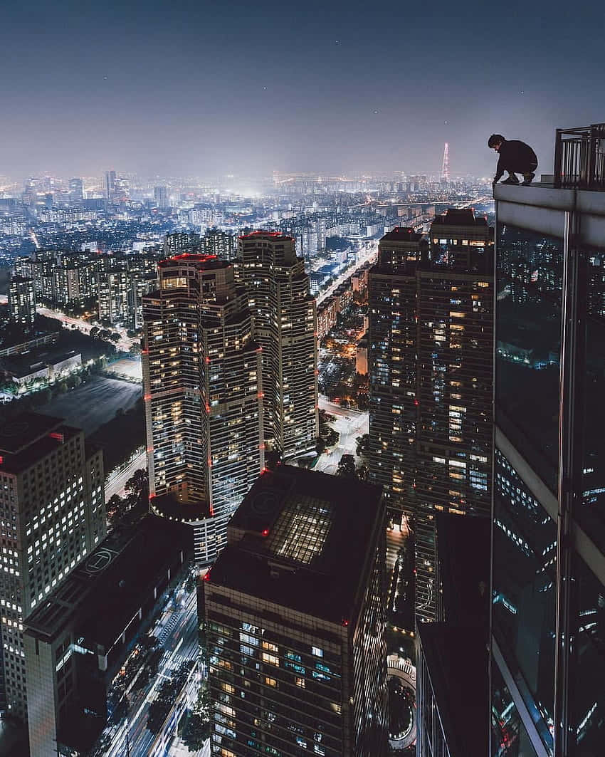 於城市頂樓享受絕美風景 壁紙 — 一個孤獨的身影在夜晚棲身於摩天大樓頂端，俯瞰著霓虹燈和星空下廣闊的城市景觀。