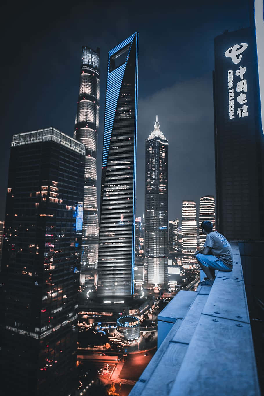 於絕美頂樓欣賞風景 壁紙 — 一個人坐在屋頂邊緣，俯瞰著閃爍窗戶的摩天大樓所構成的迷人夜景。