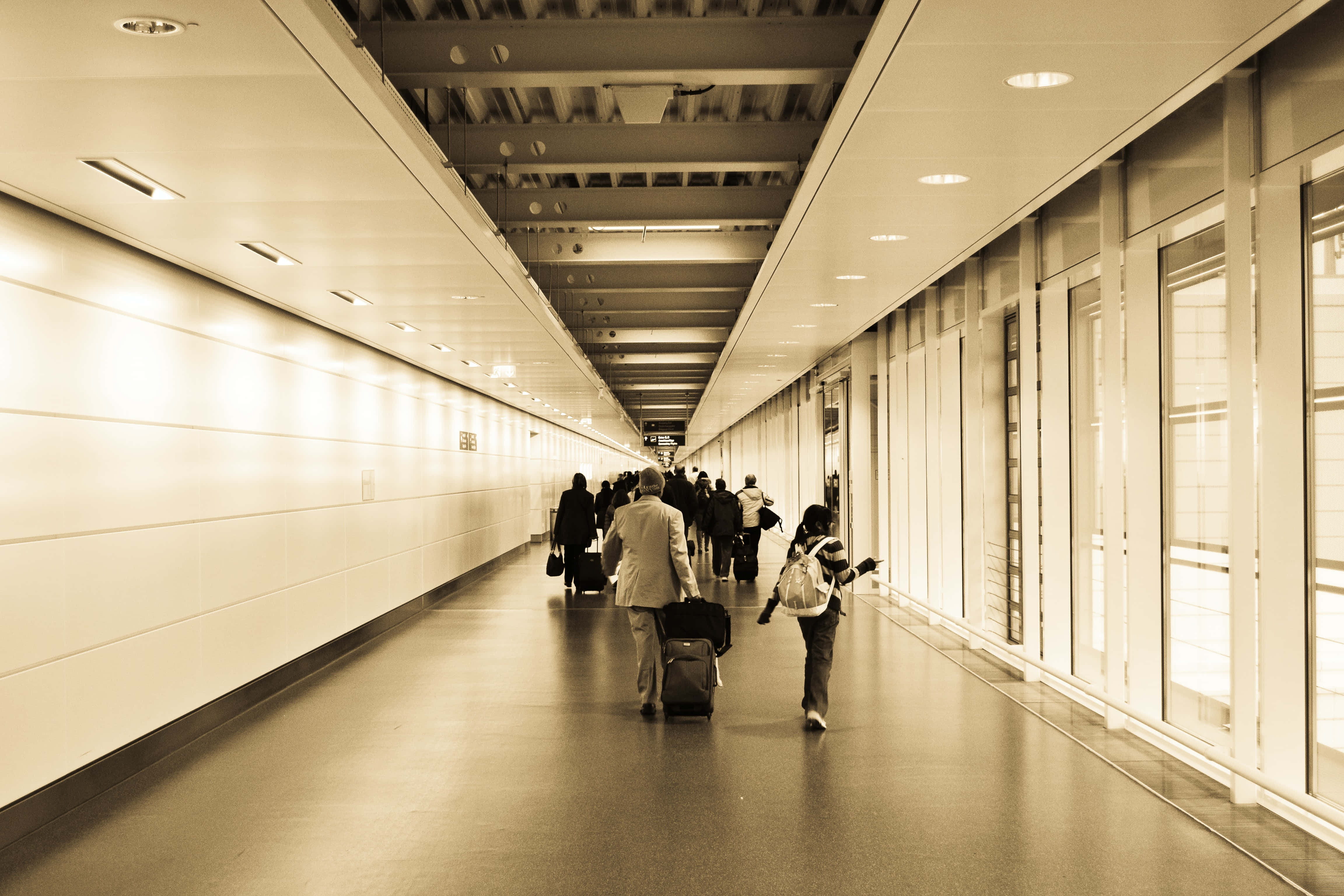 Airport 桌布 壁紙 — 旅客拖著行李箱，沿著一條長長、燈光明亮的機場走廊行走，走廊有玻璃牆和頭頂燈光。