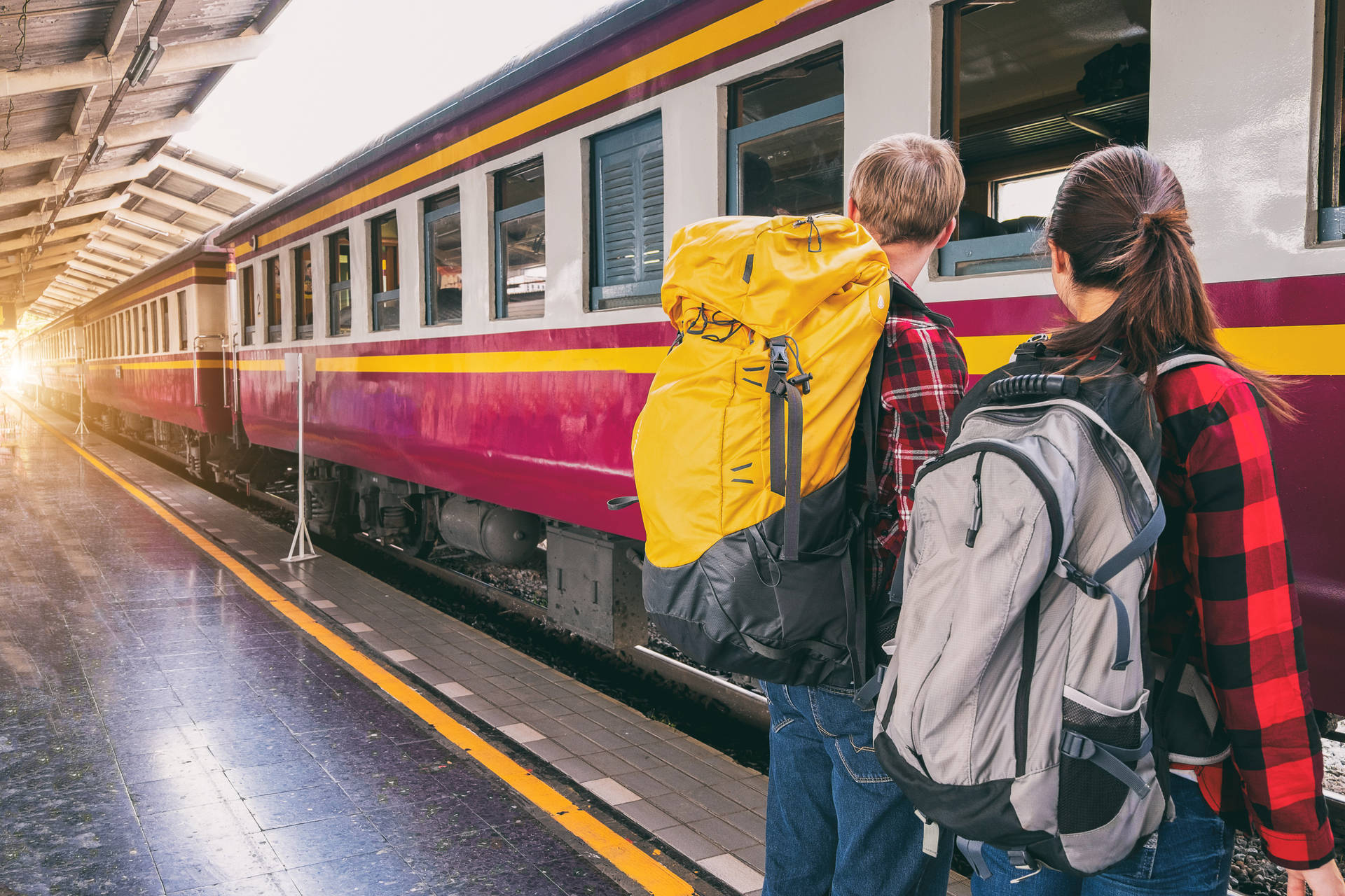 旅行4K火車之旅 壁紙 — 兩名背著背包的旅客站在火車月台上，看著一列色彩繽紛的火車抵達，金色的陽光灑滿車站。