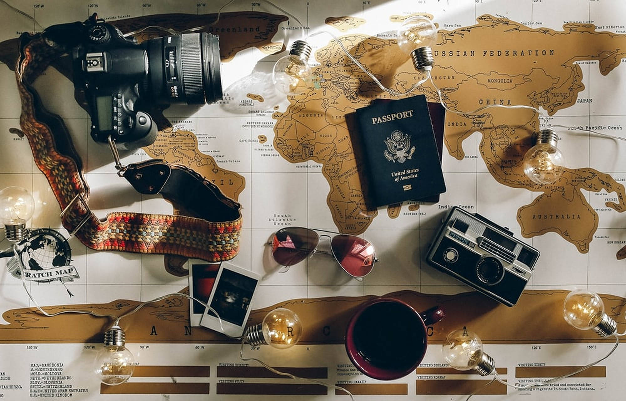 旅行必備品 壁紙 — 旅行裝備的平鋪擺設，包括一部 DSLR 相機、護照和一部復古相機，排列在世界地圖上，並配有燈串。