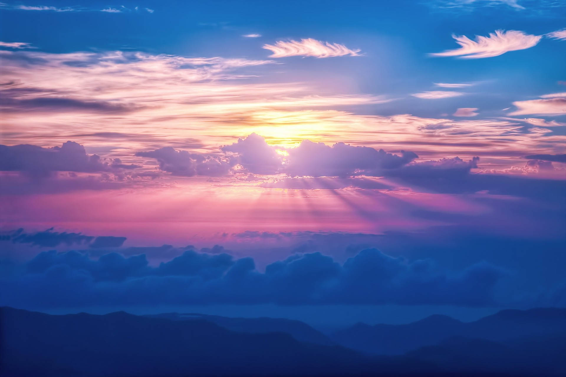 日光雲朵桌面 壁紙 — 夕陽穿透粉紅、紫色和藍色的雲層，照耀在層層疊疊的山巒輪廓之上。