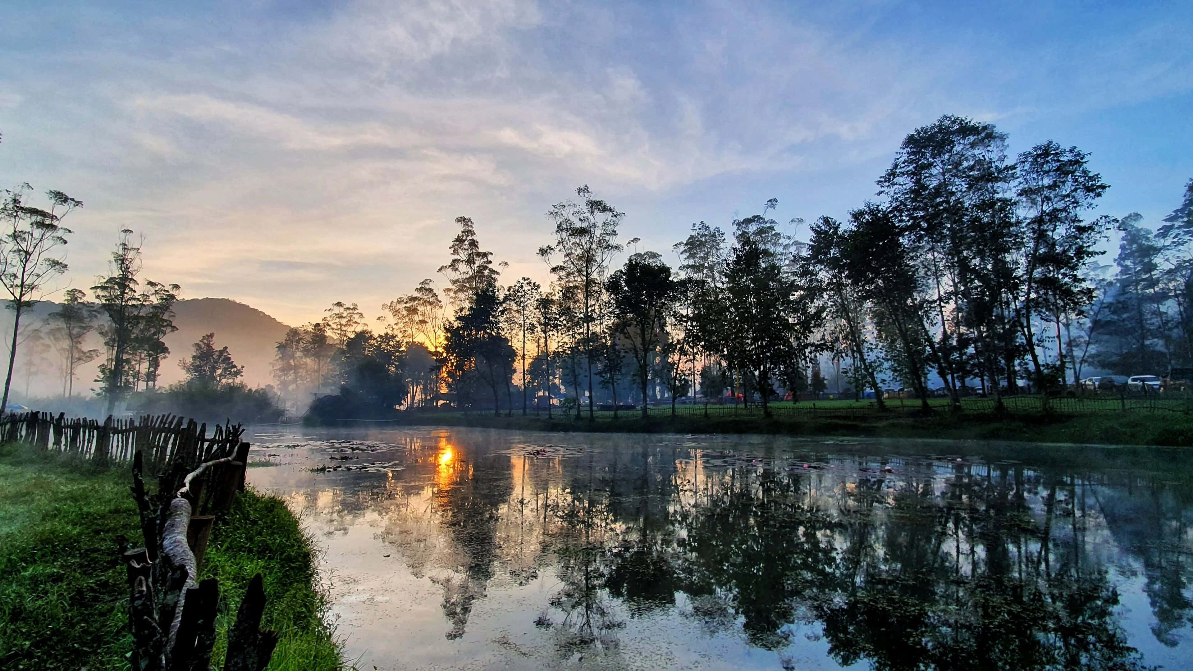 Indonesia 桌布 壁紙 — 日出在霧氣瀰漫的湖面上閃耀，映照樹木與天空，呈現寧靜的早晨氛圍。