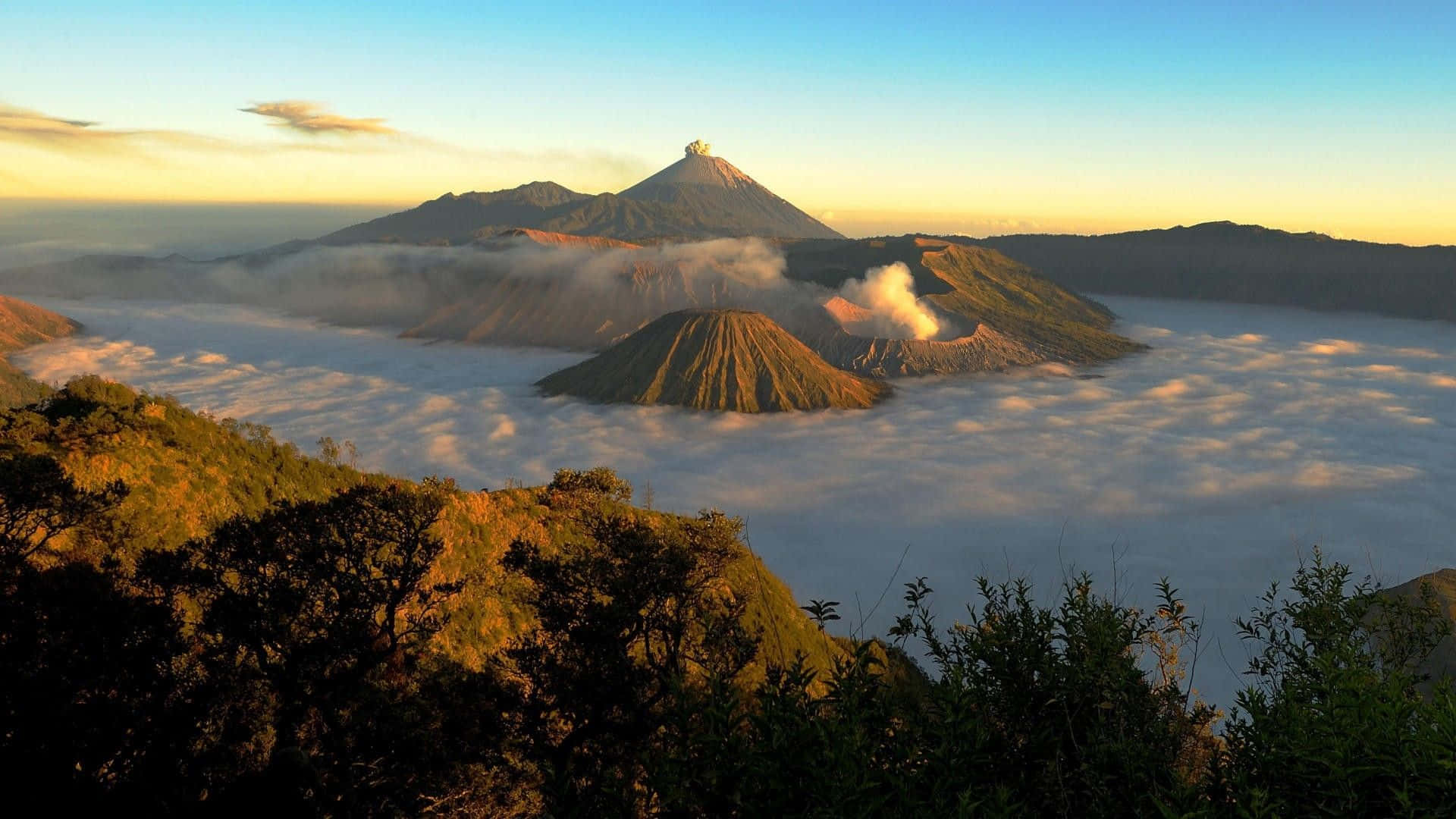 Indonesia 桌布 壁紙 — 日出時分，雄偉的火山山脈聳立在雲海之上，其中一座火山正活躍地噴出煙霧。