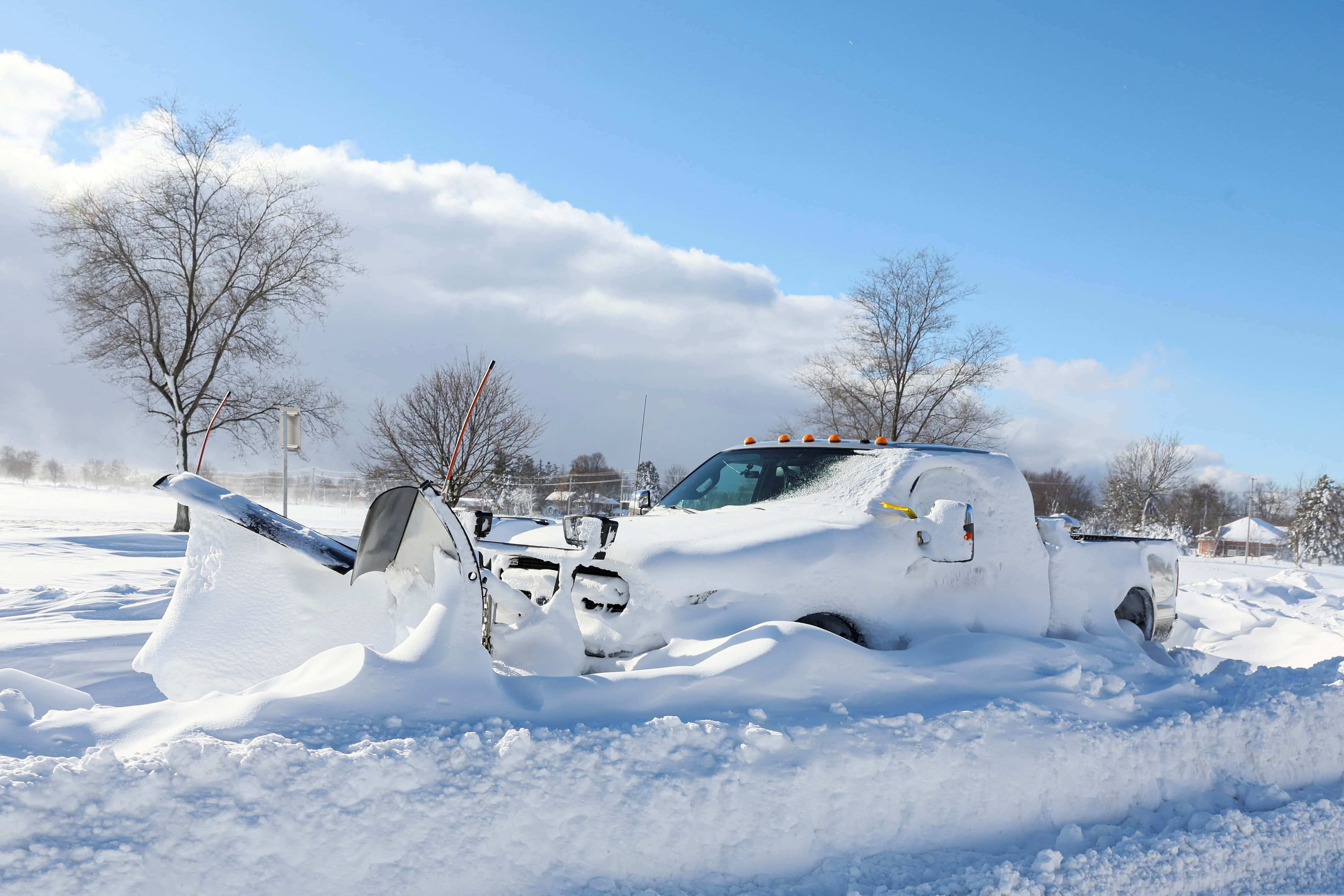 日出時分的暴雪之美 壁紙 — 一輛除雪卡車被埋在深雪中，明亮的冬季田野上有光禿禿的樹木和藍天。
